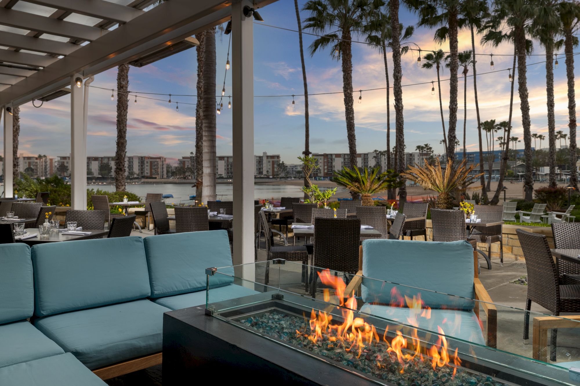 A sunny outdoor dining area with wicker chairs and teal couches around a fireplace, palm trees, string lights, and water in the background.