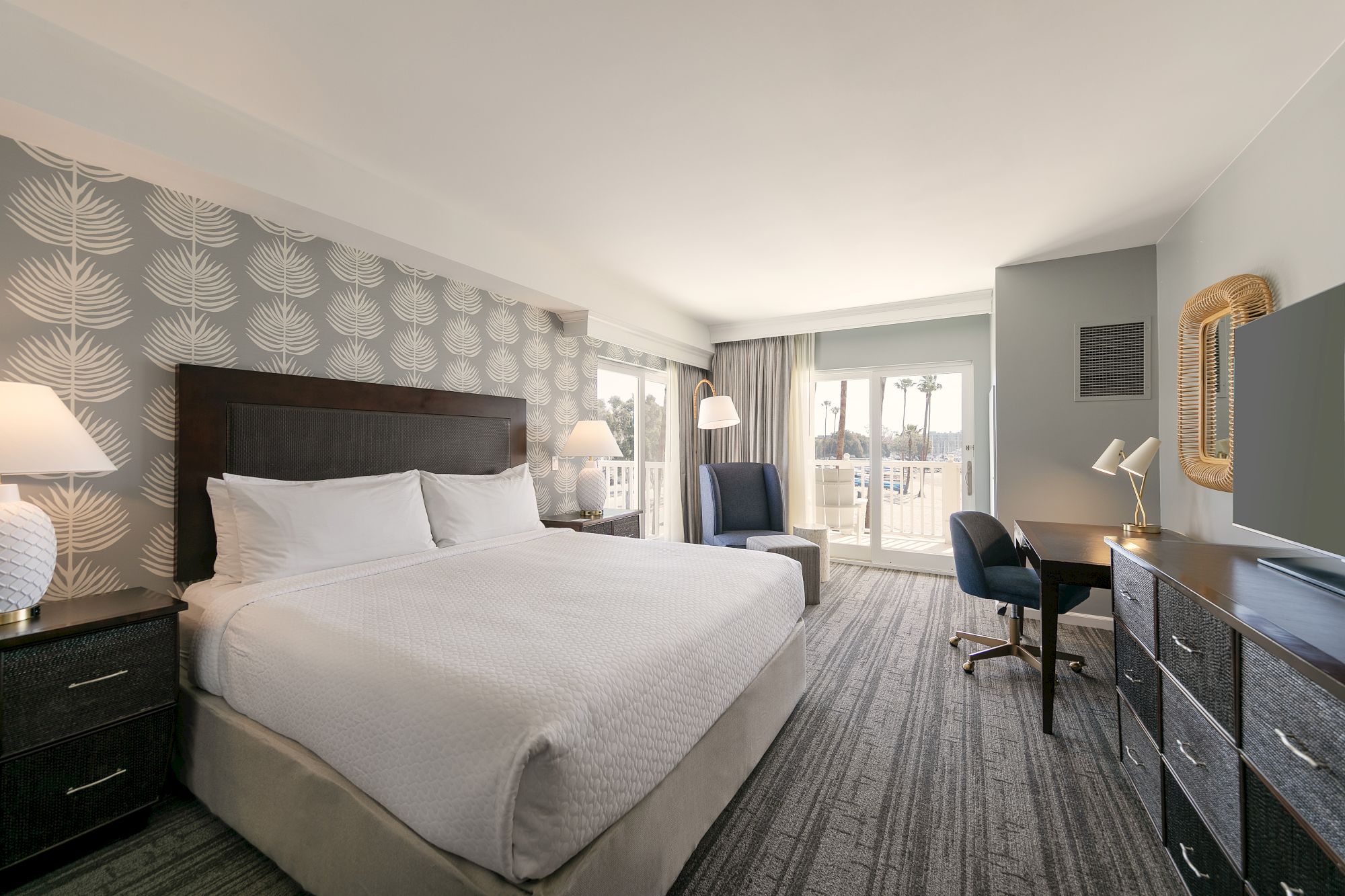 A modern hotel room with a large bed, decorative wallpaper, two nightstands, a seating area, desk with chair, and a balcony view.