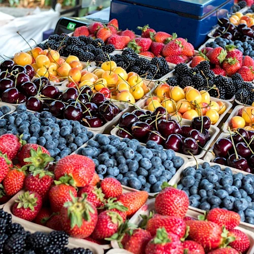 A colorful fruit market with strawberries, blueberries, cherries, blackberries, and peaches arranged in trays at a stall. Ending the sentence.