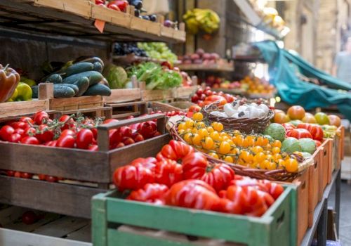 A vibrant outdoor market with crates of red tomatoes, yellow cherry tomatoes, cucumbers, peppers, and other fresh produce arranged on wooden stalls.
