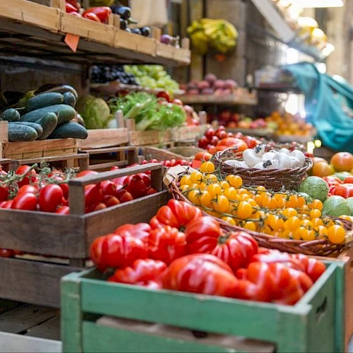 A vibrant outdoor market with crates of red tomatoes, yellow cherry tomatoes, cucumbers, peppers, and other fresh produce arranged on wooden stalls.