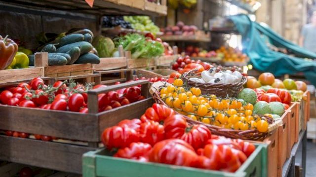 A vibrant outdoor market with crates of red tomatoes, yellow cherry tomatoes, cucumbers, peppers, and other fresh produce arranged on wooden stalls.