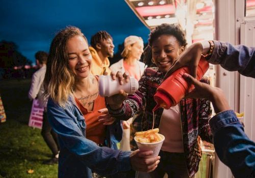 A group of friends sharing funnel cake and drinks at a fair, smiling and having fun together, with one pouring into cups.