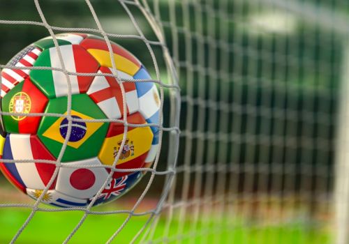 A close-up soccer ball, covered in international flags, stuck in a goal net on a grassy field.
