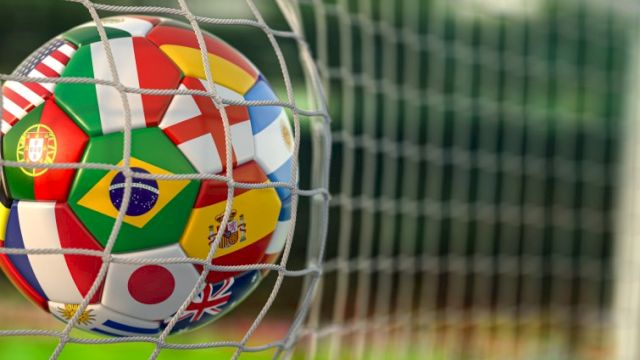 A close-up soccer ball, covered in international flags, stuck in a goal net on a grassy field.