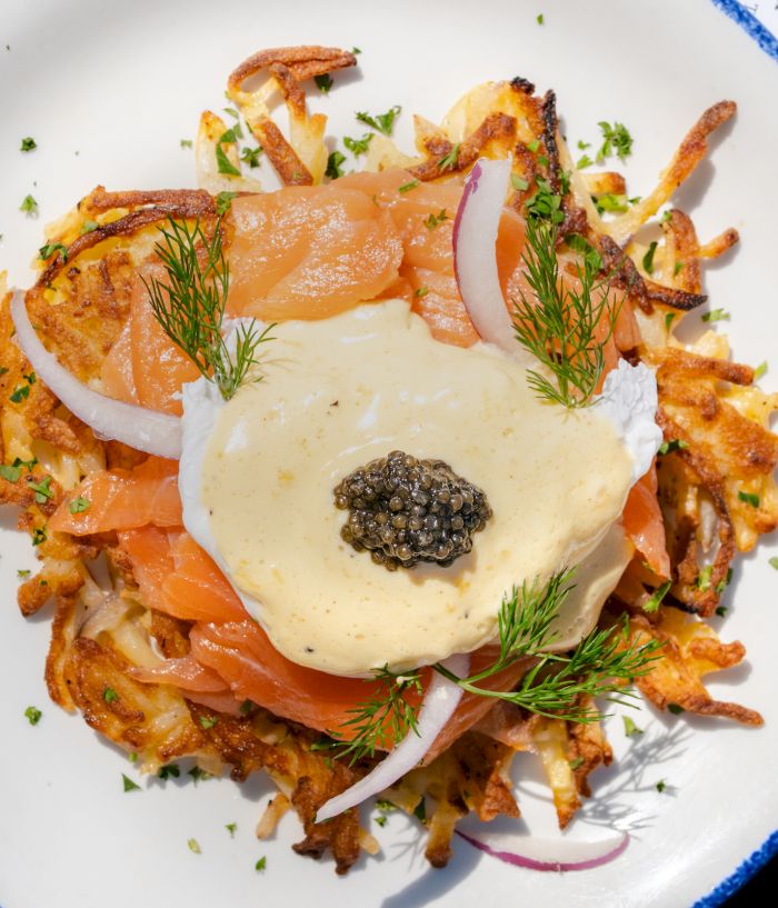 A plate of crispy potatoes topped with smoked salmon, onion slices, dill, a creamy sauce, and a dollop of caviar on top.