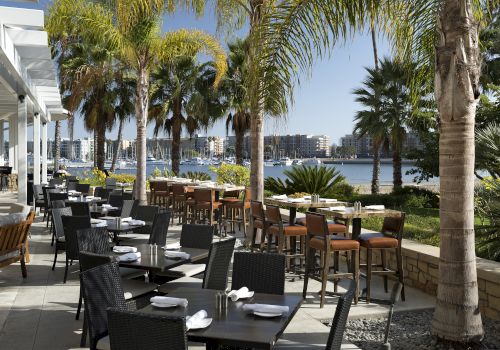 A seaside restaurant patio with black tables and chairs, palm trees, and a water view under a sunny sky, outdoor dining setup.