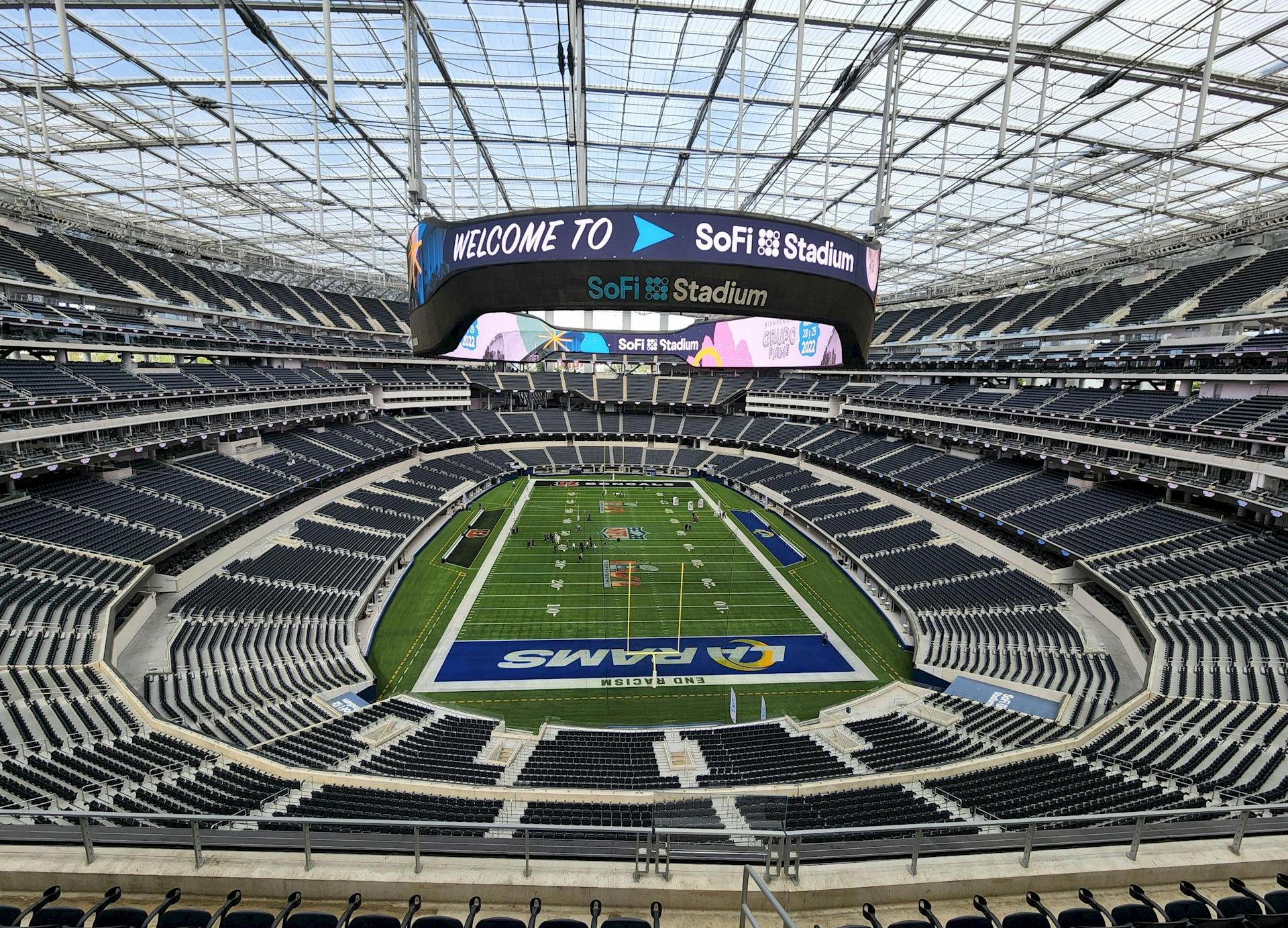 Inside a large NFL stadium with a green football field, empty seats, and a jumbotron displaying &ldquo;WELCOME TO&rdquo; and &ldquo;SOFi Stadium&rdquo; ending with a period.