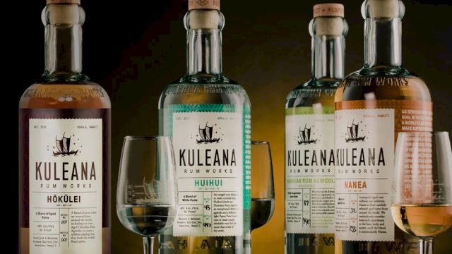 A row of Kuleana whiskey bottles with glasses, dark background, labels visible, three flavors.