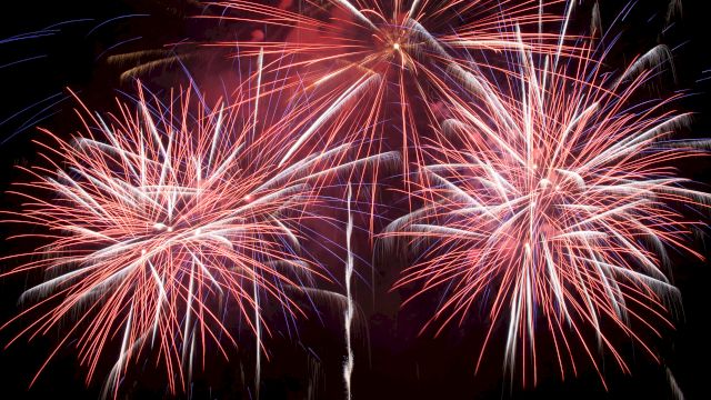 Colorful red, blue, and white fireworks burst in a dark night sky, forming multiple starburst trails.