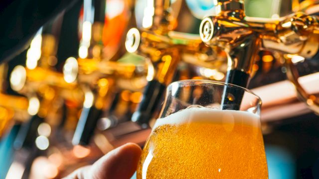 A hand holds a foamy beer in front of shiny beer taps, with warm golden lights in a bar.