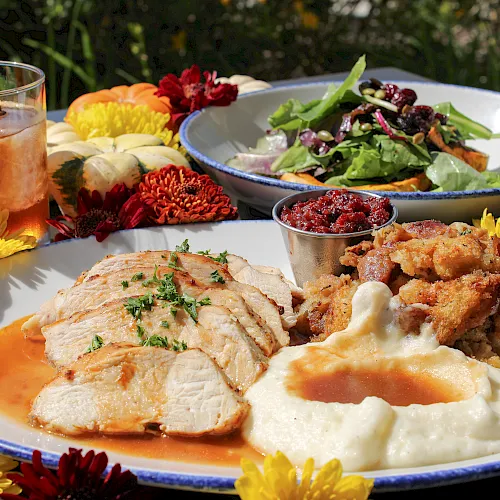 Outdoor feast spread: sliced meat with creamy sauce, roasted meat, green salad and olives, plus a glass drink and flowers nearby.