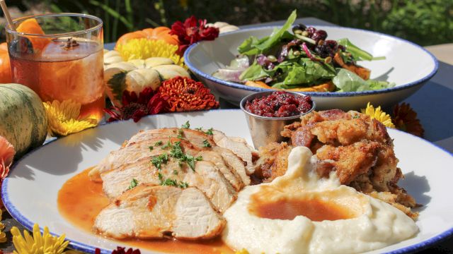 Outdoor feast spread: sliced meat with creamy sauce, roasted meat, green salad and olives, plus a glass drink and flowers nearby.